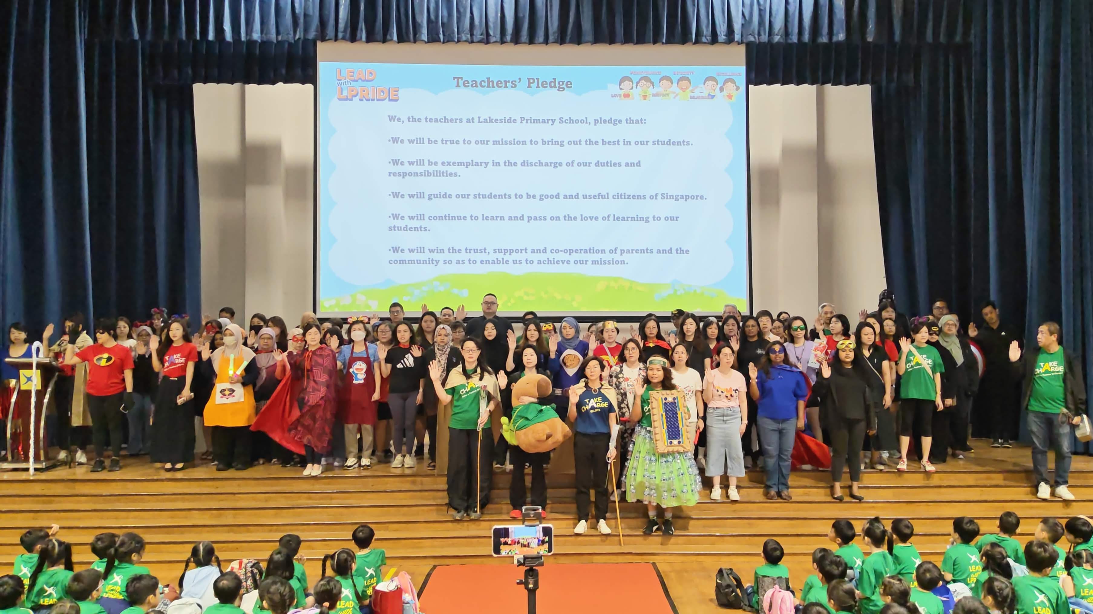A large group of teachers in costumes stand on a school hall stage with their right hands raised, reciting the Teachers' Pledge displayed on a projector screen behind them. Students in green T-shirts are seated in the foreground watching the ceremony.